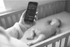 A person is holding a smartphone showing a baby monitor app next to a sleeping baby in a crib. Produktbild-Vorschau