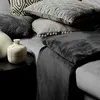 A close-up view of several pillows and a blanket stacked on a sofa, with two decorative vases on a side table. Produktbild-Vorschau