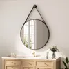 A round mirror with a black strap hanging over a vanity with a faucet and some decorative items. Voir les images du produit