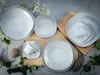 A collection of white square-shaped porcelain dishes and cups are arranged on a textured gray surface with a bamboo mat and some greenery. Produktbild-Vorschau