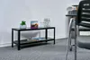 A black metal shelving unit with two shelves, placed against a white wall on a grey carpeted floor. There are a few items on the top shelf, including a potted plant, a calendar, and some bottles of water. Some magazines are on the bottom shelf. A chair is  Produktbild-Vorschau