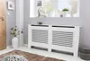 A white radiator cover with horizontal slats is placed in a room with a window and a vase of flowers. Podgląd zdjęcia produktu