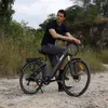 A man is sitting on an electric bicycle on a dirt path with vegetation in the background. Produktbild-Vorschau