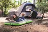 A couple camping in a forest setting. A woman is relaxing on an air mattress reading a book, while a man is setting up a tent. Podgląd zdjęcia produktu