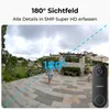 A person walks on a paved patio in front of a house, with a security camera visible in the foreground. Produktbild-Vorschau