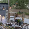 An outdoor dining table setting with string lights overhead and decorative spheres. Produktbild-Vorschau