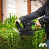 A person is using an electric hedge trimmer to trim a green hedge in front of a house. Náhľad obrázku produktu