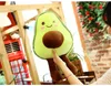 A person holding a plush avocado toy in a room with wooden structures and some plants. Produktbild-Vorschau