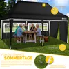 A family is enjoying a meal under a black pop-up gazebo in a grassy backyard. Produktbild-Vorschau