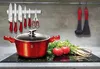 A red pot with a glass lid sits on a black stovetop, surrounded by fresh vegetables and kitchen utensils hanging on a magnetic strip. Produktbild-Vorschau