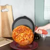 A cast iron pan with a freshly baked pizza is being held by a hand with a red silicone grip. A wooden cutting board and a red plate are also visible. Produktbild-Vorschau