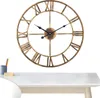 A large metal wall clock with Roman numerals is displayed against a white background, with a desk and some stationery in the foreground. Produktbild-Vorschau