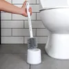 A hand holding a white and gray toilet brush over its white holder, next to a white toilet. The background is tiled white. Podgląd zdjęcia produktu