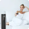 A woman sitting on a white sofa holding a remote control, with a black tower fan next to her. Podgląd zdjęcia produktu