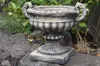 A stone urn sits on rocks in a garden setting, with greenery in the background. Produktbild-Vorschau