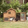 A wooden chicken coop with an attached run, situated in a garden setting with lush greenery and a brick building in the background. Two chickens, one white and one black, are visible in and around the coop. Produktbild-Vorschau