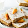 Several slices of carrot cake with white icing and marzipan carrot decorations are arranged on a white plate. A glass of milk is visible in the background. Produktbild-Vorschau