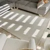 A rug is placed on a wooden floor in front of a sofa and an armchair, with sunlight casting shadows on the rug. Voir les images du produit