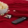 A bowl of popcorn, a snack packet, and a remote control are placed on a red blanket, suggesting a cozy movie-watching setup. Produktbild-Vorschau