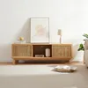 A wooden TV stand with slatted doors, placed in a living room setting with a framed picture and a lamp. Podgląd zdjęcia produktu