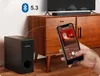 A hand holding a smartphone is shown in front of a wooden cabinet with a TV and soundbar on top, and a subwoofer to the left. The phone displays a music app. Produktbild-Vorschau