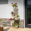 A multi-tiered metal plant stand with potted flowers is displayed outdoors against a textured wall and stone base. Náhľad obrázku produktu