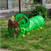 A young girl is playing inside a green caterpillar-shaped tunnel in a grassy outdoor area. Produktbild-Vorschau
