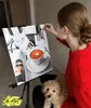 A woman is painting a "paint by numbers" picture of a cat and coffee, with her dog sitting beside her. Produktbild-Vorschau