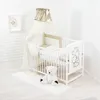 A baby crib set with bedding, a canopy, and a teddy bear on a rug, set against a white brick wall. Produktbild-Vorschau