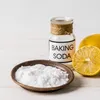 A small glass jar of baking soda, a small bowl of baking soda, and a sliced lemon on a white wooden surface. Produktbild-Vorschau