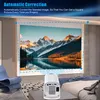 A projector is shown in a bedroom setting, projecting an image of mountains and a lake onto a wall. The projector is small and white, placed on a table. Produktbild-Vorschau
