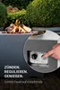A close-up of a modern outdoor fire pit with a hand pressing a button to ignite the flame, surrounded by lush greenery. Produktbild-Vorschau