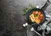 A frying pan with cooked salmon, tomatoes, mushrooms, and rosemary on a dark, textured surface. Náhľad obrázku produktu