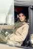 A woman sits in the driver's seat of a vehicle, looking out the window. Produktbild-Vorschau