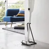 A white vacuum cleaner is standing upright on a black stand in a room with a blue sofa and a rug in the background. Produktbild-Vorschau