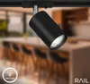 A black spotlight fixture is mounted on a track light system, with a blurred kitchen or dining area in the background. Anteprima dell’immagine prodotto
