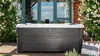A hot tub is situated outdoors in a garden setting, surrounded by flowering bushes and next to a building with large windows. Produktbild-Vorschau