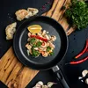 A close-up overhead shot of a black frying pan filled with cooked shrimp, garnished with parsley, a slice of lemon, and a red chili pepper. The pan is placed on a wooden cutting board, with bread, garlic, and more chili peppers scattered around the dark ba Produktbild-Vorschau