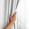 A hand is parting a white blackout curtain to reveal a sheer curtain and a window with greenery outside. Produktbild-Vorschau