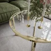 A close-up view of a round glass coffee table with a gold frame, placed on a carpet next to a green sofa. Two white candles in gold candleholders and a bouquet of flowers are on the table. Náhľad obrázku produktu