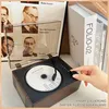 A hand is shown adjusting a knob on a vintage-style Bluetooth speaker with a CD player, with books in the background. Produktbild-Vorschau