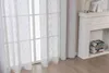 A close-up view of white linen-look curtains hanging in front of a window, with a white fireplace and stacked books visible to the right. Produktbild-Vorschau