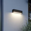 A black rectangular LED wall light mounted on a light gray textured wall, emitting a warm white light downwards. Produktbild-Vorschau