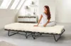 A woman is sitting on a foldable bed in a room with slanted ceilings and skylights. Anteprima dell’immagine prodotto