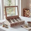 A wooden cutlery box with a glass lid is placed on a white table, filled with cutlery. In the background, a window shows a garden, and a stack of firewood is visible. Podgląd zdjęcia produktu