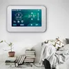 A digital thermostat is mounted on a white wall above a sofa with decorative pillows and a blanket, and a stack of books and a potted plant are on the floor below. Náhľad obrázku produktu