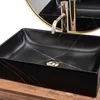 A black rectangular basin with gold marbling, a gold faucet, and a gold drain sits on a wooden surface. A round gold-rimmed mirror is visible in the background. Produktbild-Vorschau