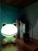 A frog-shaped LED night light sits on a wooden desk next to a stack of books and a small lantern. Náhľad obrázku produktu
