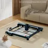 A laser engraving machine is placed on a rug in a living room setting, next to a sofa and a small table with a vase of dried flowers. Náhled obrázku produktu