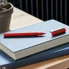 A red pen with a silver cap rests on a dark grey notebook, which is stacked on another dark grey notebook. A potted succulent is visible to the left. Produktbild-Vorschau